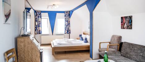 A room with a bed, a sofa, a table, and a chair. There is a window with curtains, a radiator, and a picture on the wall.