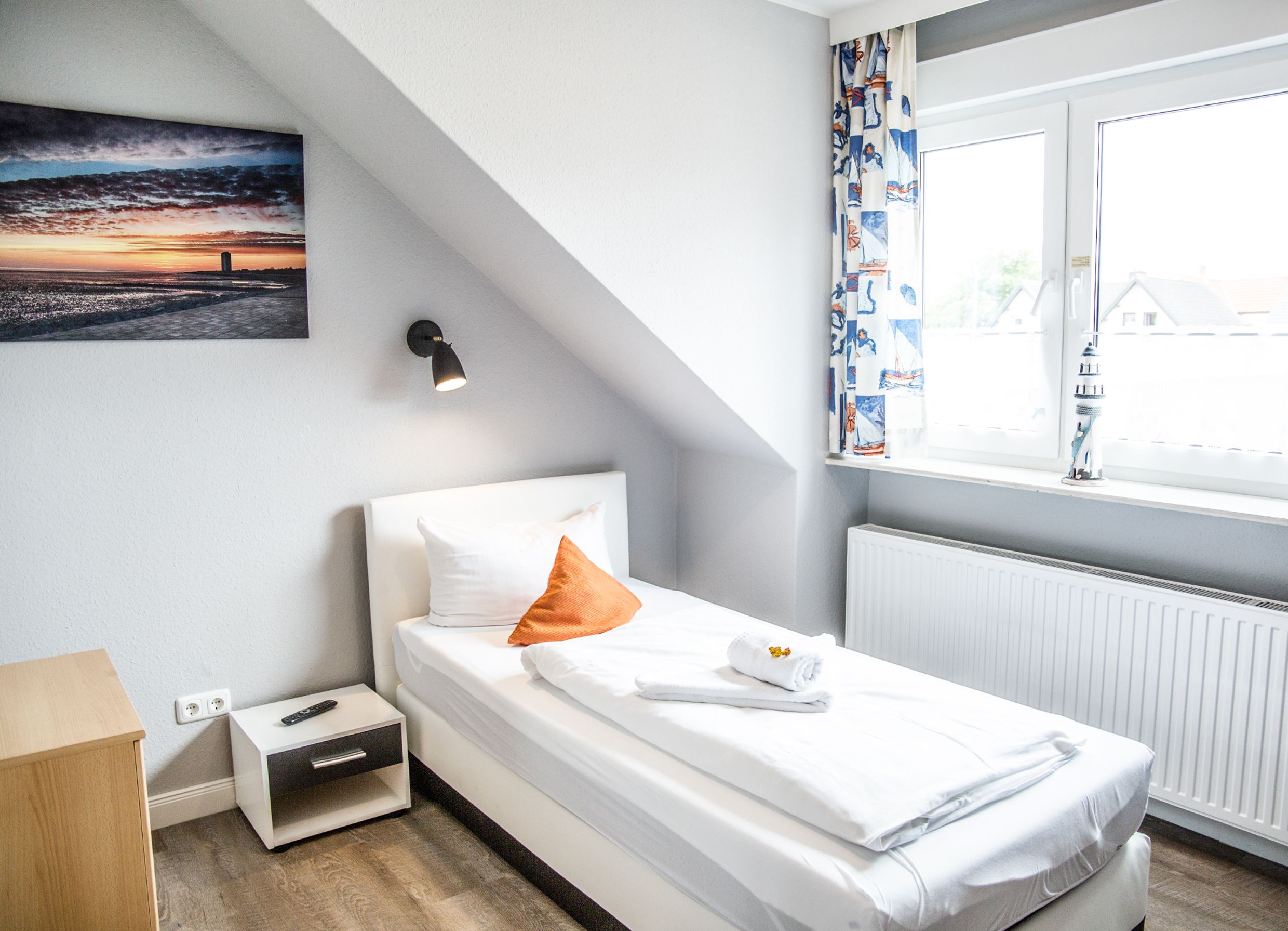 A neatly arranged bedroom with a bed, a bedside table, a nightstand, a wall-mounted lamp, a window with curtains, and a painting on the wall.