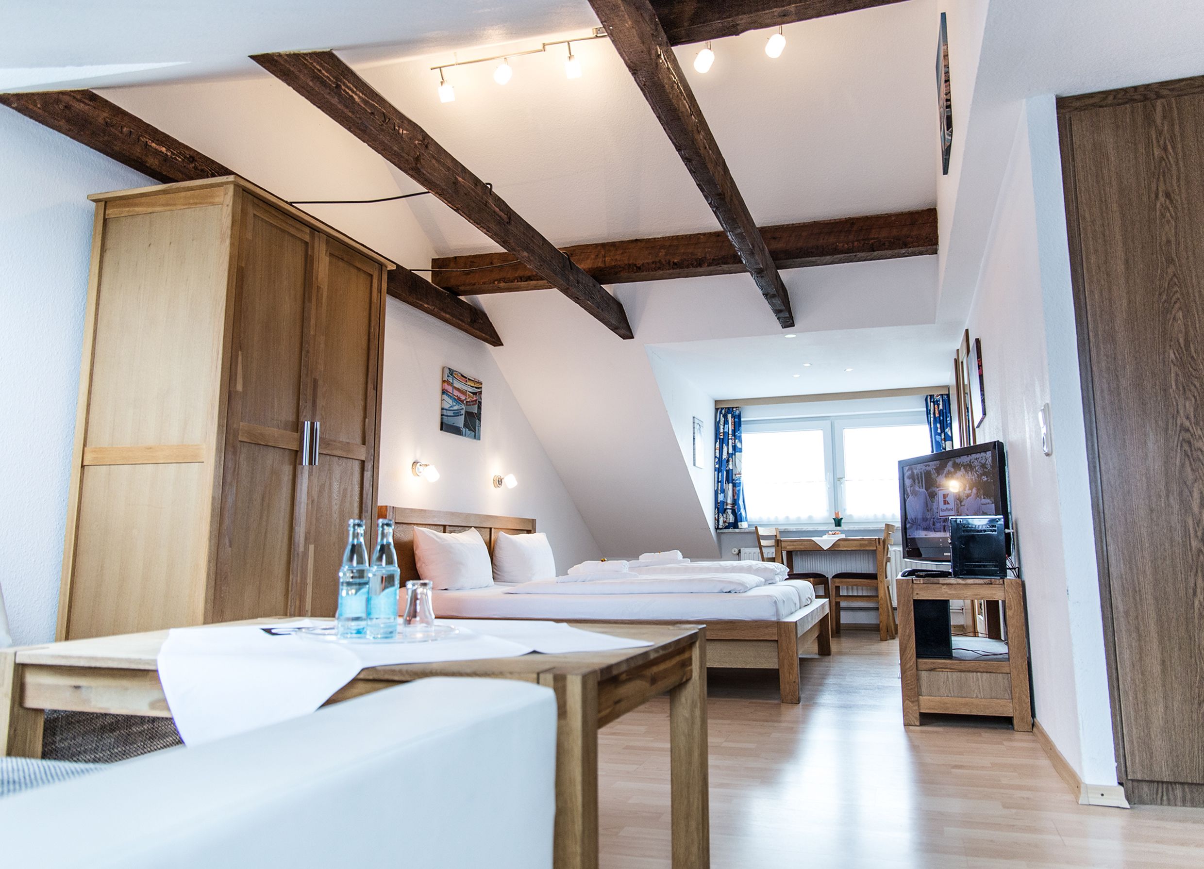 A modern, well-lit room with a sloped ceiling, wooden beams, a large wardrobe, a dining table with chairs, and a television.