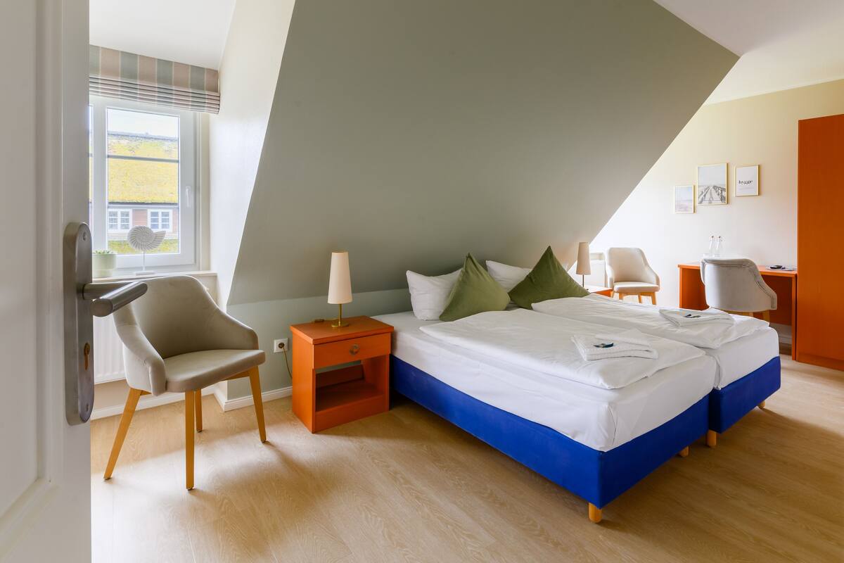 Un dormitorio con techo inclinado, una cama con ropa de cama blanca y azul, dos sillas, una mesita de noche con una lámpara y una ventana con vistas a un edificio.