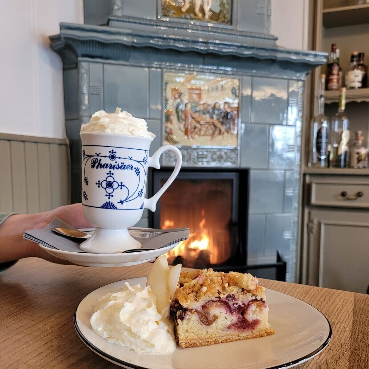 Sobremesa com chantilly e uma fatia de bolo em um prato, uma xícara com a palavra 'Pharisier' escrita e uma lareira ao fundo.