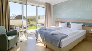 Habitación de hotel con una cama grande, una silla, una mesa con vasos y una botella, y un balcón con vistas al exterior.