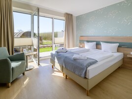 Habitación de hotel con una cama grande, una silla, una mesa con vasos y una botella, y un balcón con vistas al exterior.