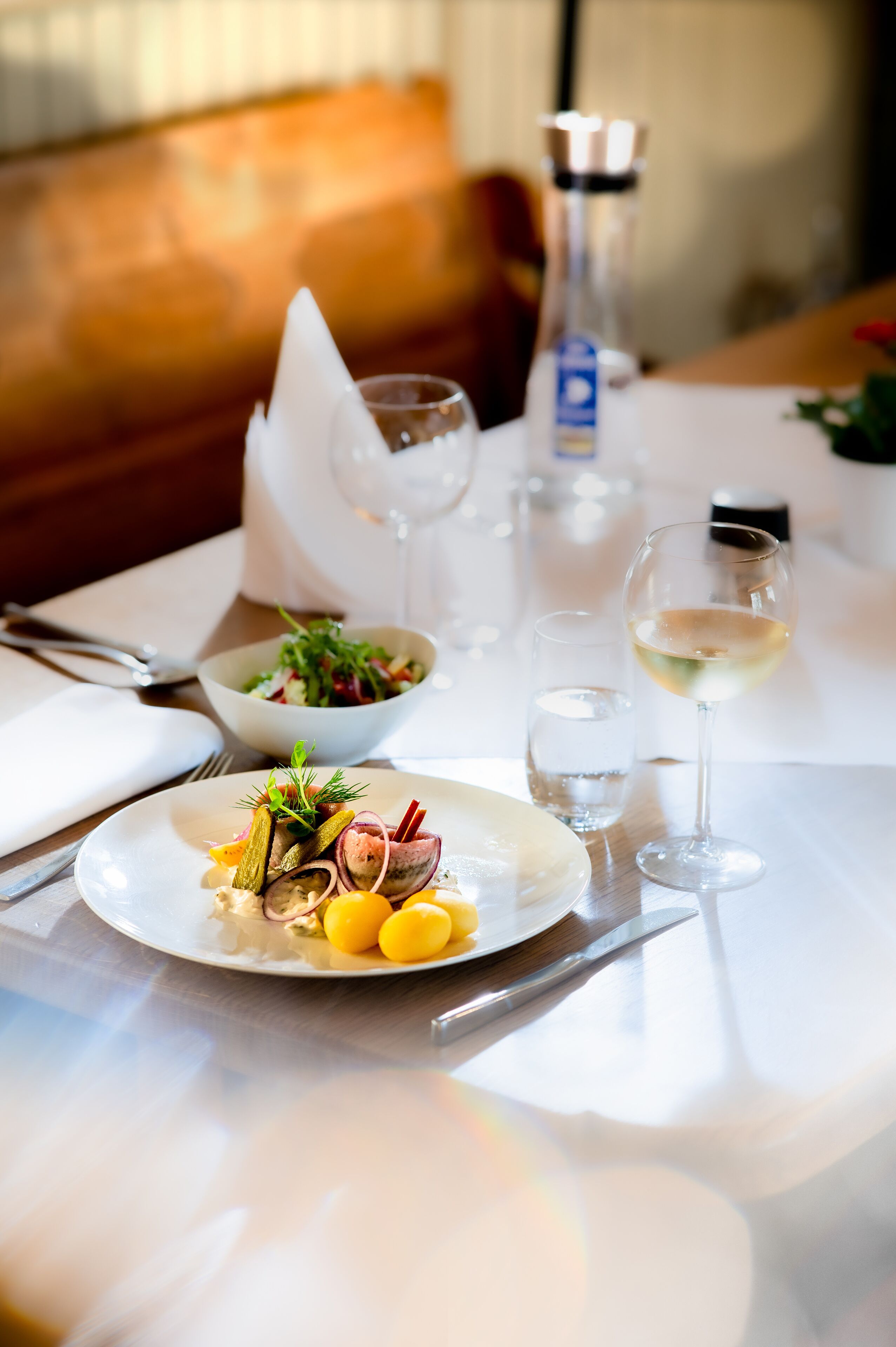 Una mesa de comedor con mantel blanco, un tazón de ensalada, un plato con pepinillos y huevos, una copa de vino blanco y un salero.