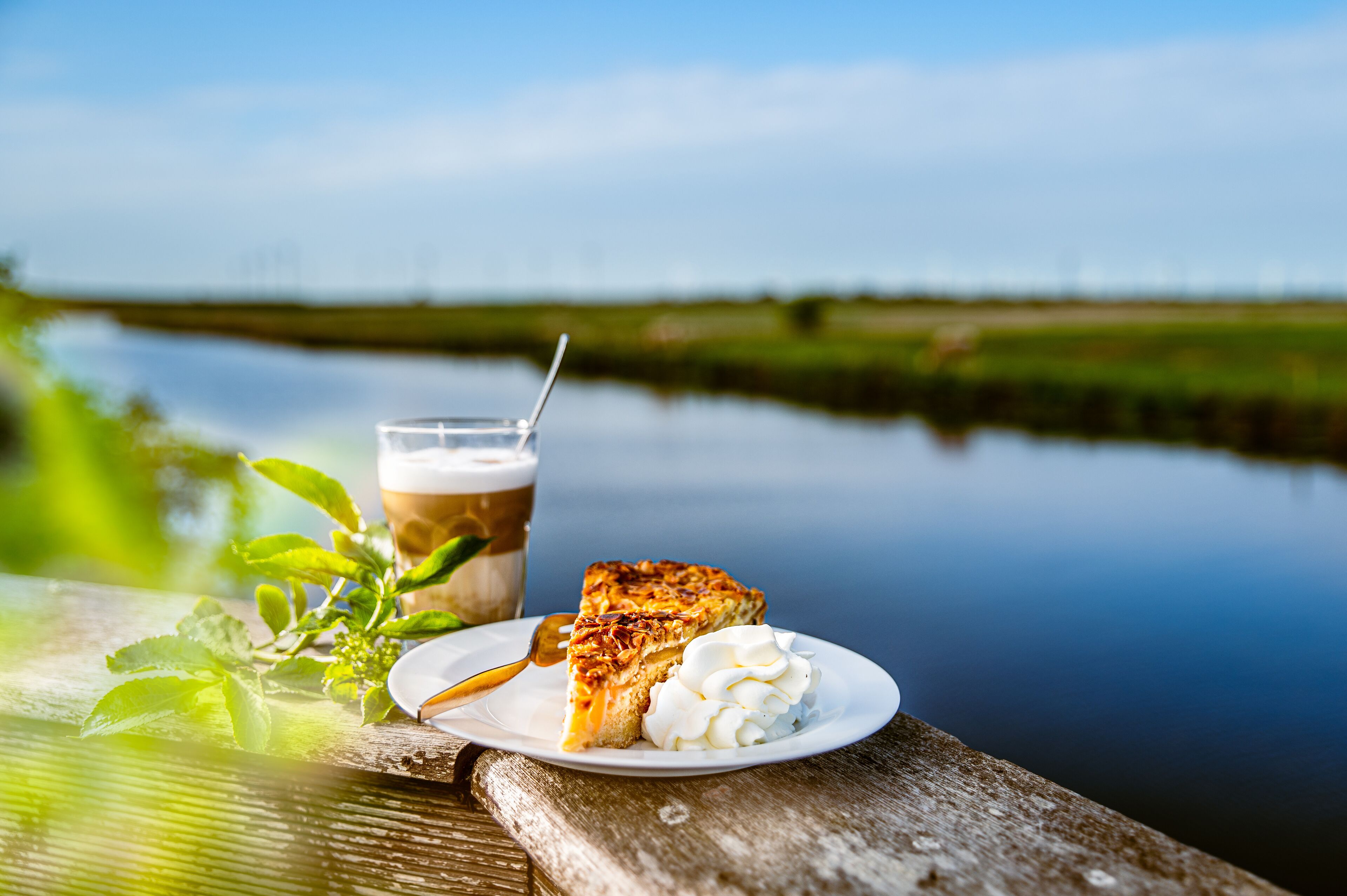 Une part de tarte garnie de crème fouettée, posée sur une assiette, un verre de café glacé et une cuillère, le tout disposé sur une table en bois, près d’une étendue d’eau calme.