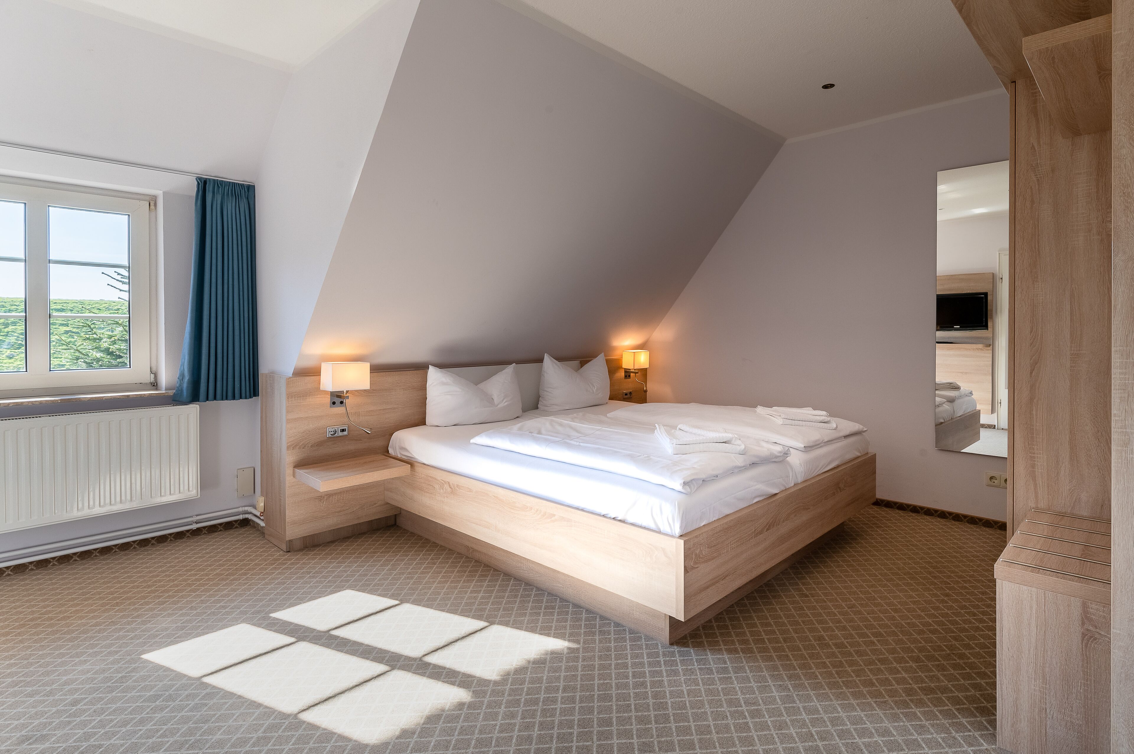 Een moderne slaapkamer met een schuine kap, een groot bed met witte lakens, twee bedlampen, een houten nachtkastje en uitzicht op het buitenlands landschap door een raam met blauwe gordijnen.