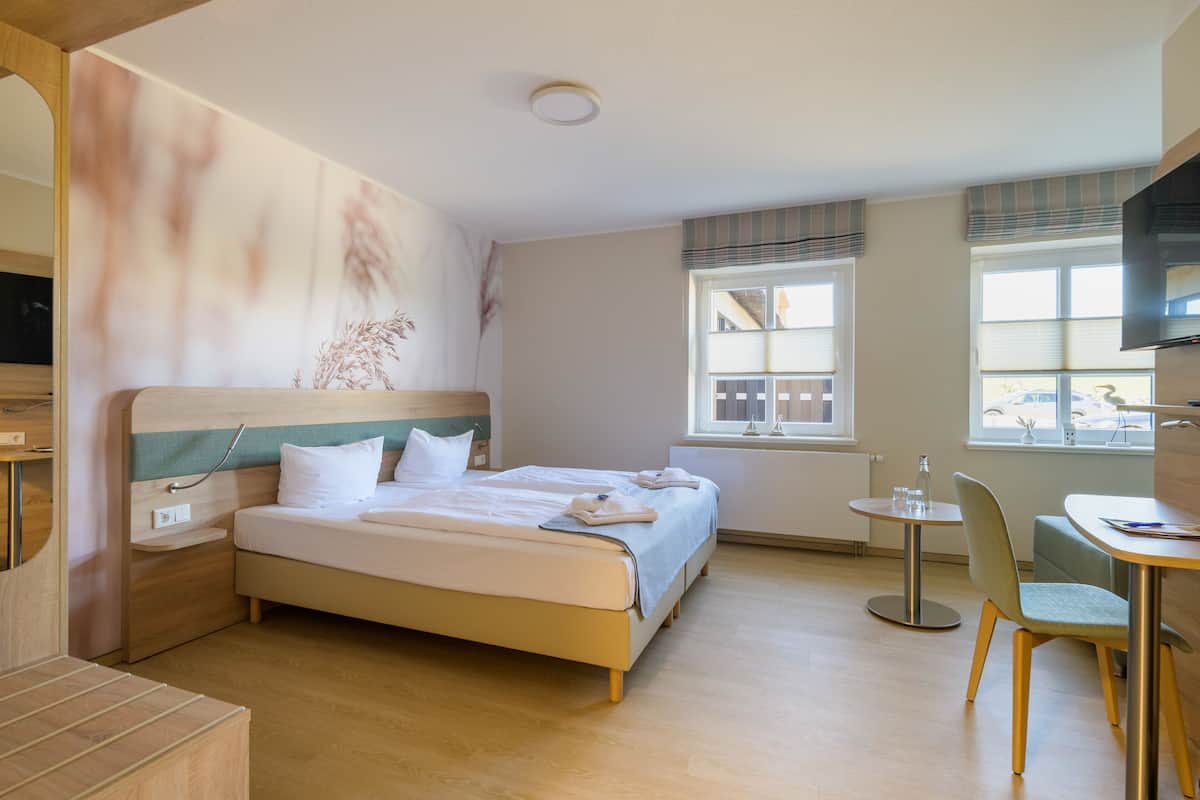Habitación de hotel con cama, mesita de noche, silla y una pequeña mesa con otra silla.