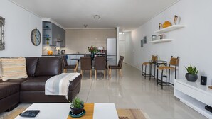 Uma sala de estar moderna com um sofá de couro marrom, uma área de jantar com mesa de vidro e uma cozinha ao fundo.