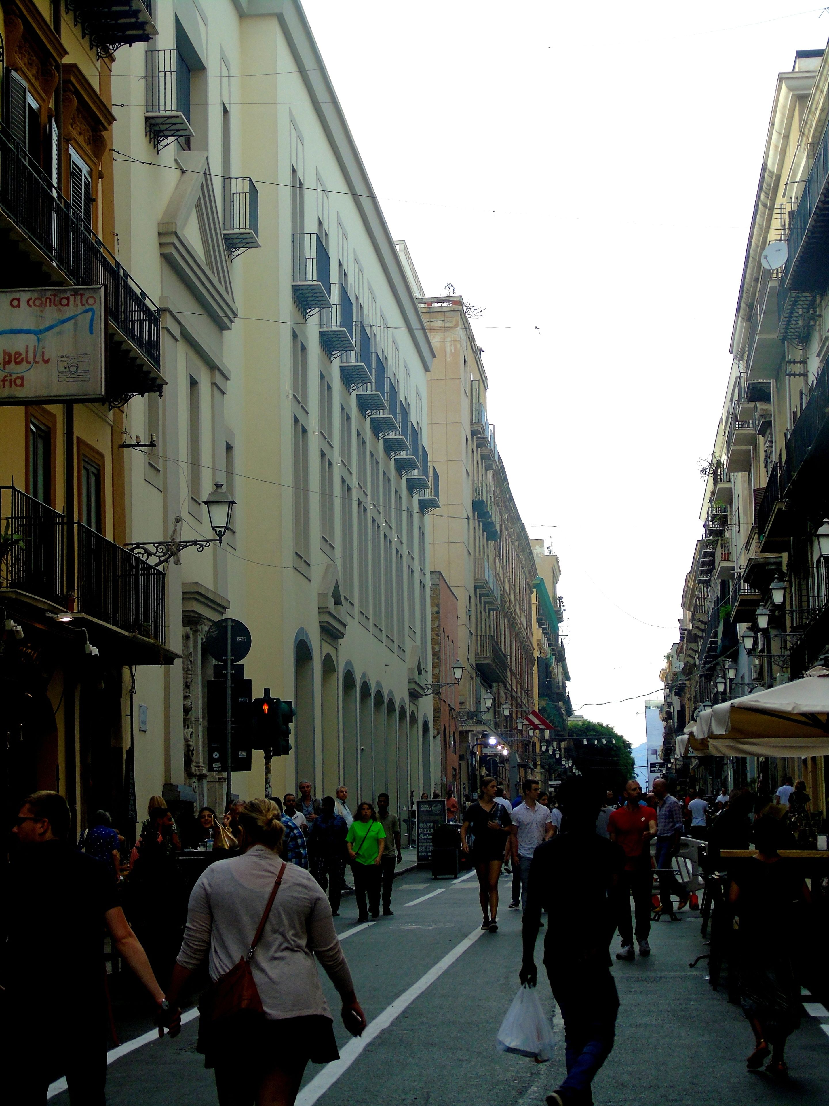 A crowded street with buildings on either side, featuring balconies and street lamps.