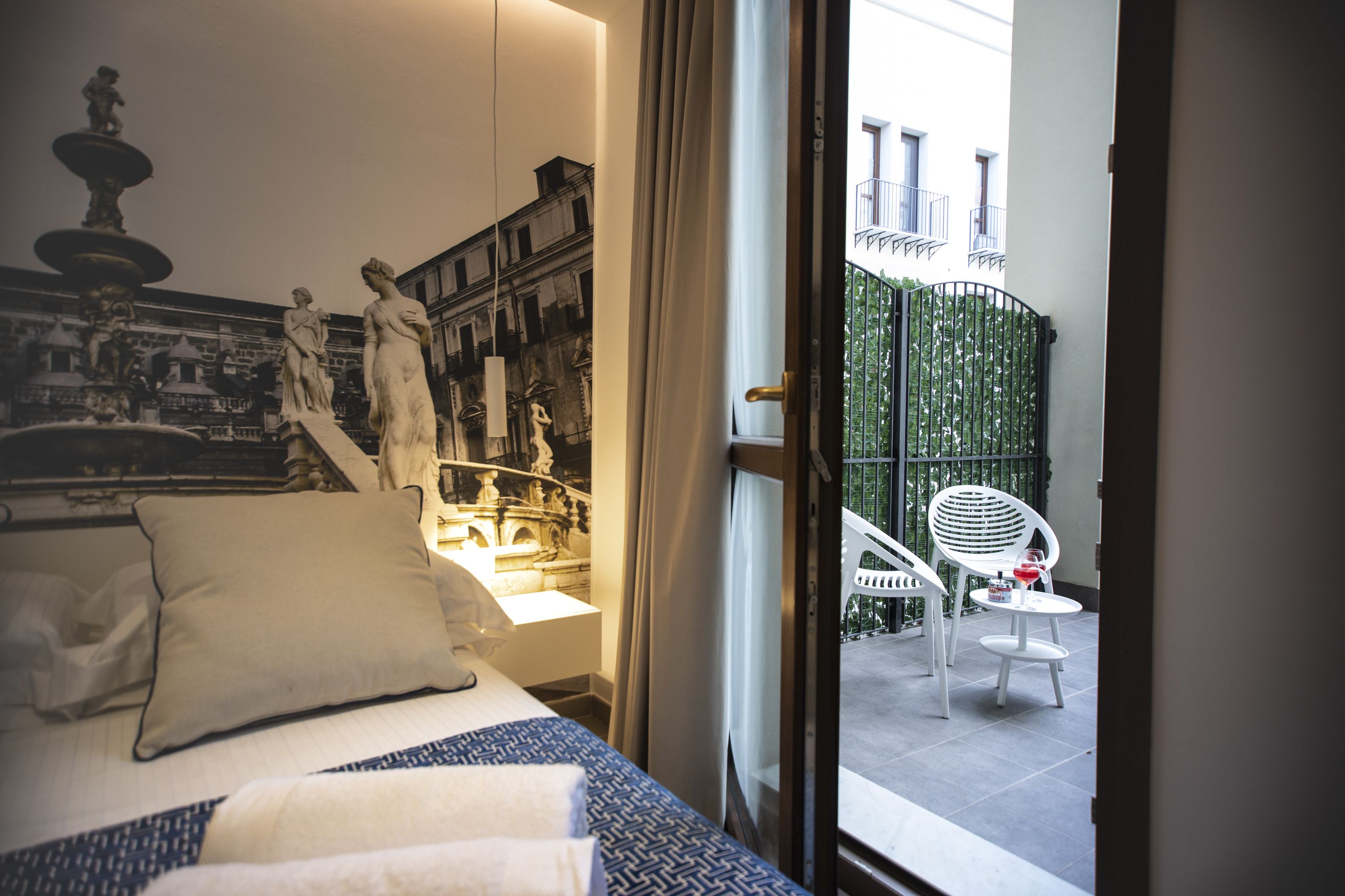 A hotel room with a bed, a view of a balcony with a chair and table, and a mural of a fountain and statues.
