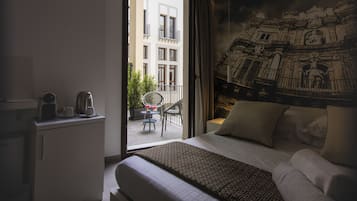 A hotel room with a bed, a desk with a coffee maker, a chair, and a balcony with a view of a building and greenery.