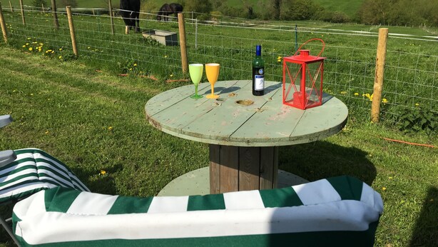 Una mesa redonda de madera con un mantel verde, dos copas de vino, una botella y un farol rojo sobre un campo cubierto de hierba con una valla.