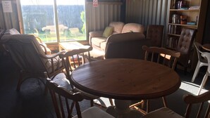 Un rincón interior acogedor con una mesa redonda de madera y sillas, una estantería y vistas al exterior.