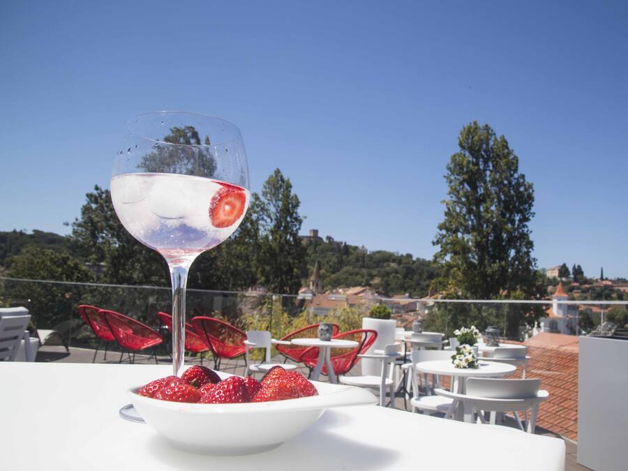 A table with a glass of a clear beverage, a bowl of strawberries, and red chairs in an outdoor setting with a view of trees and buildings.