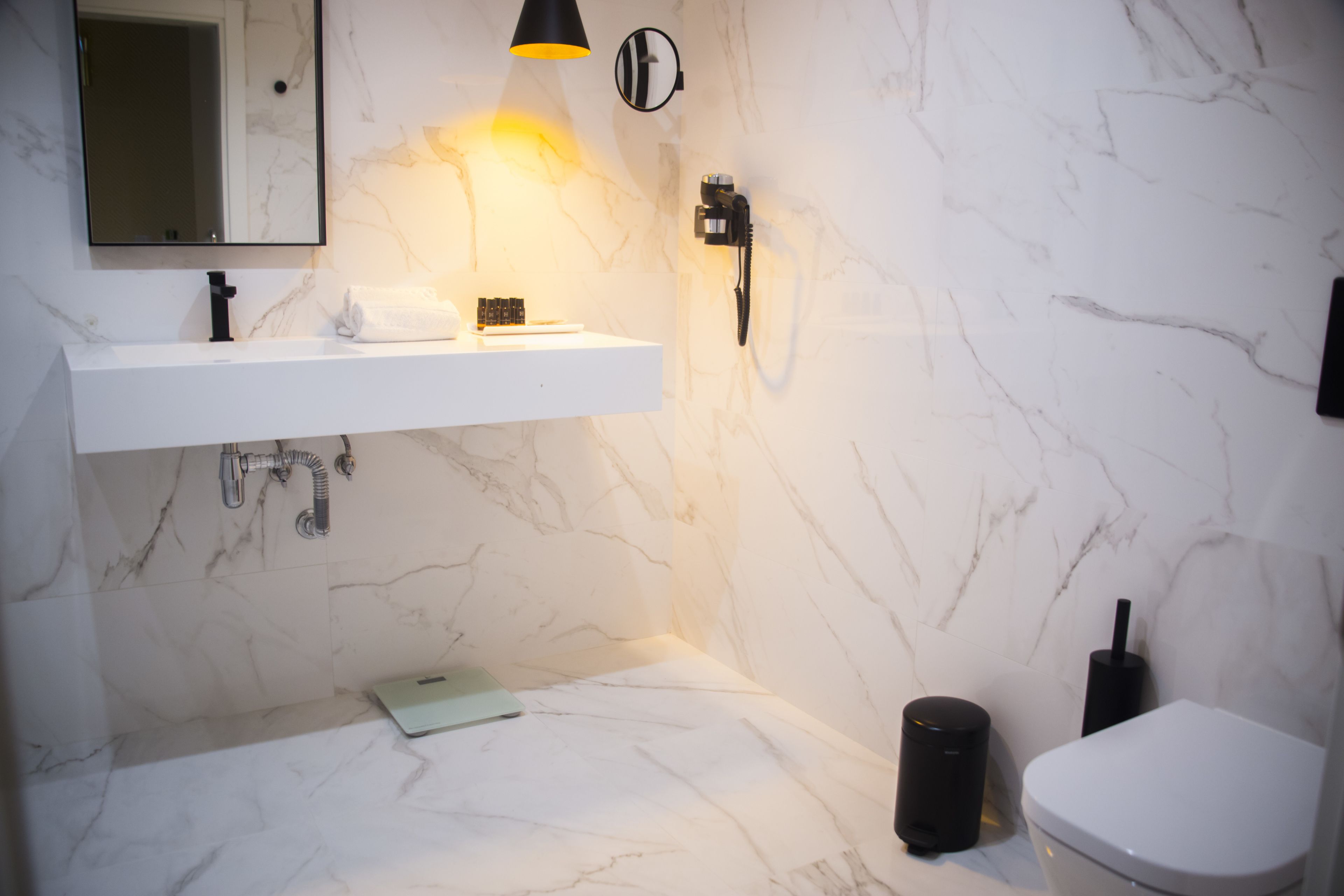 A modern bathroom with a white marble sink, a mirror, a towel, a handheld showerhead, a toilet, and a trash bin.