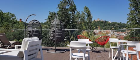 Een terras met witte loungestoelen en tafels, een hangstoel en uitzicht op een heuvel met gebouwen.