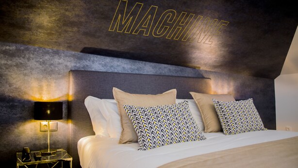 A modern hotel room with a large bed, patterned pillows, a nightstand with a lamp, and a wall with the word "MACHINE" illuminated. - Thomar Boutique Hotel (Tomar)