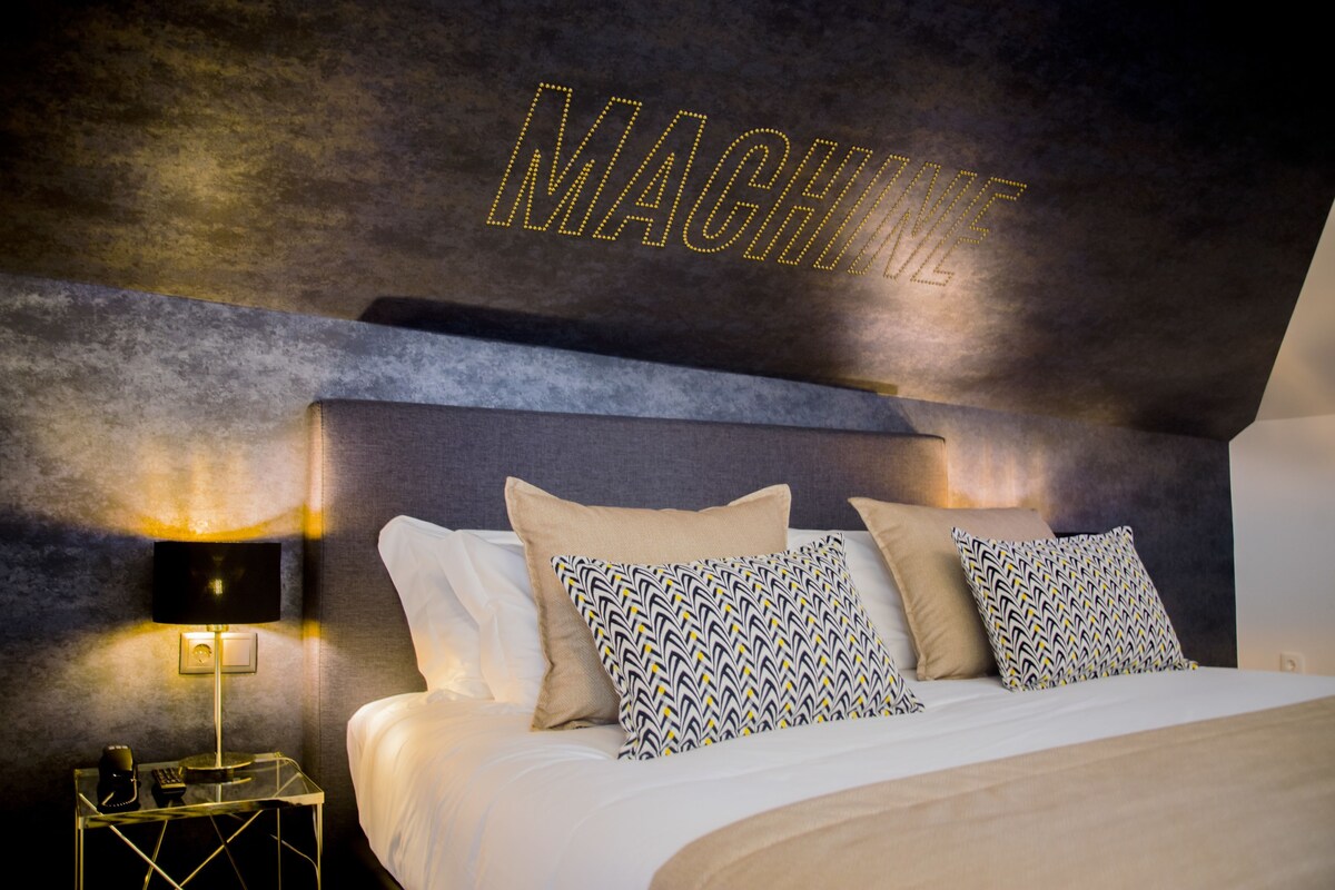 A modern hotel room with a bed, pillows, a nightstand, a lamp, and a wall with the word 'MACHINE' illuminated.