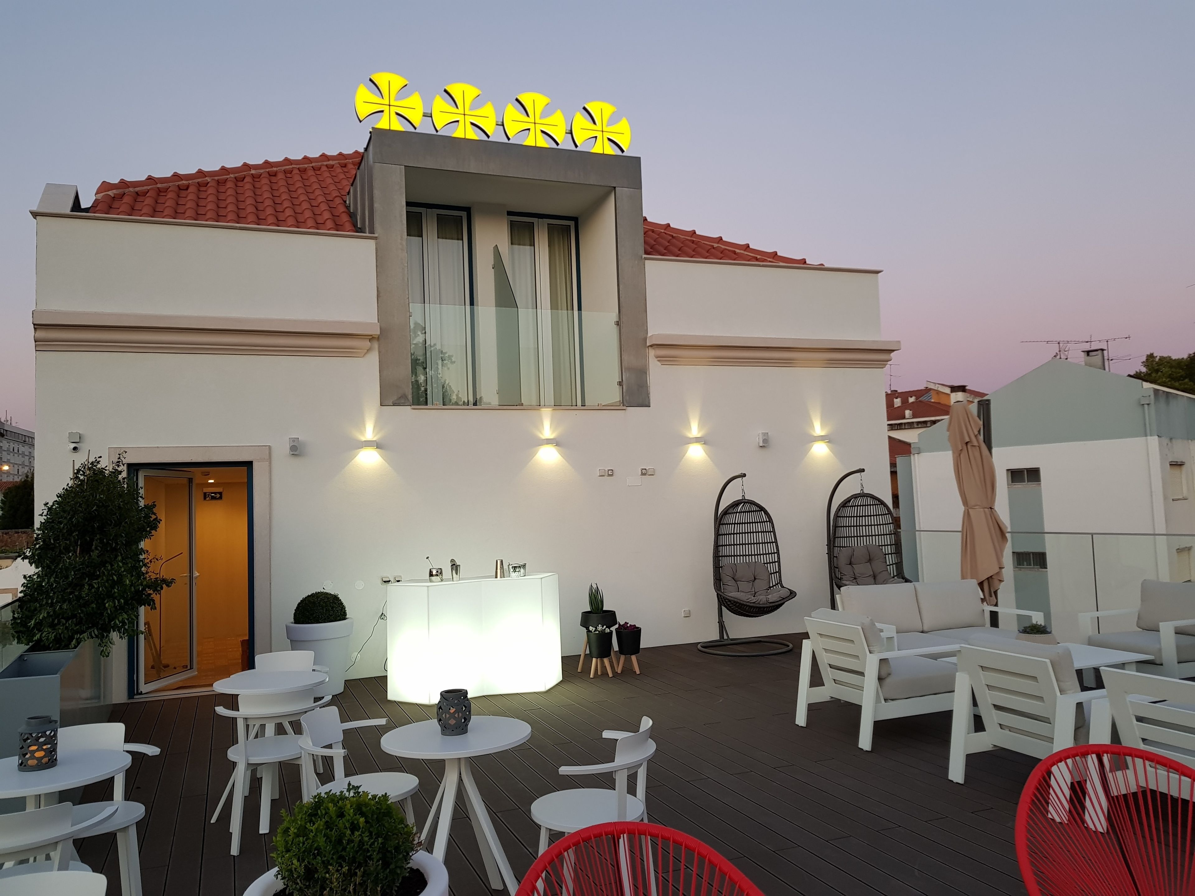A modern building with a white facade, a red-tiled roof, and a yellow sign. There are outdoor seating areas with white and red chairs, and a bar area with a white counter and decorative items.