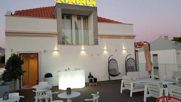 A modern building with a white facade, a red-tiled roof, and a yellow sign. There are outdoor seating areas with white and red chairs, and a bar area with a white counter and decorative items.