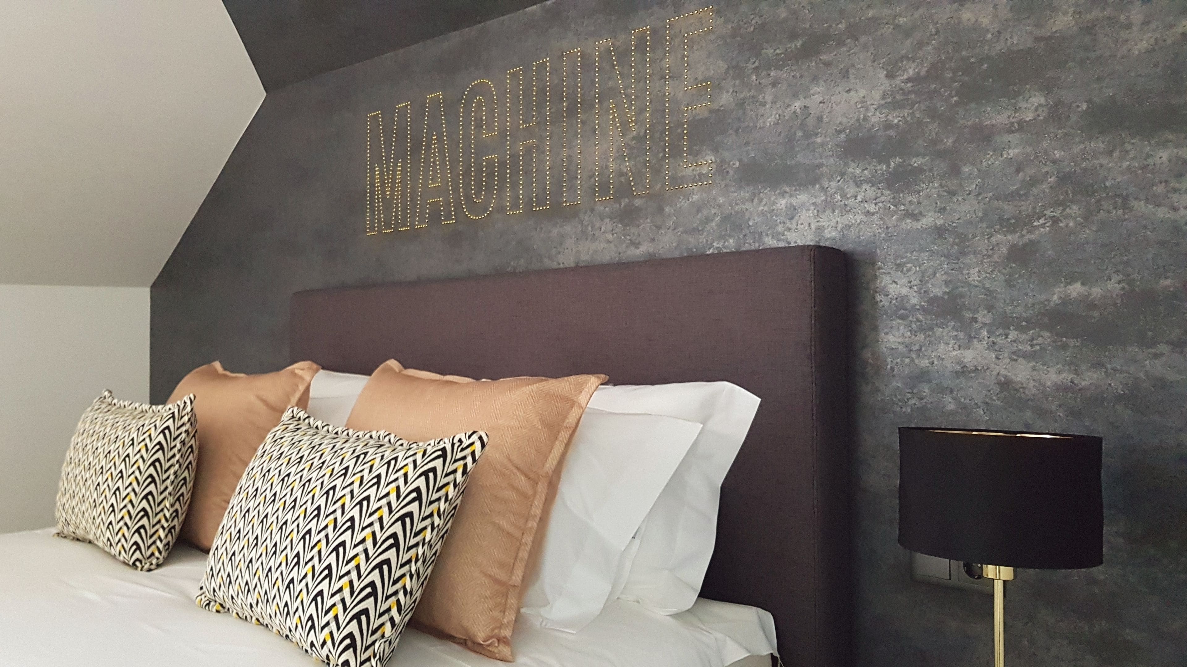 A modern bedroom with a dark headboard, patterned pillows, and a wall-mounted light fixture.