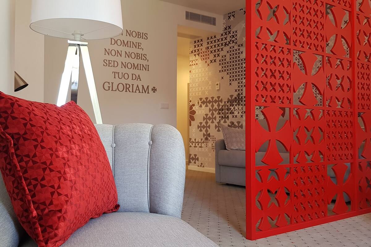 Ein modernes Wohnzimmer mit einem grauen Sofa, einem roten geometrischen Kissen, einer weißen Stehlampe und einem dekorativen roten Raumteiler mit Ausschnitten.