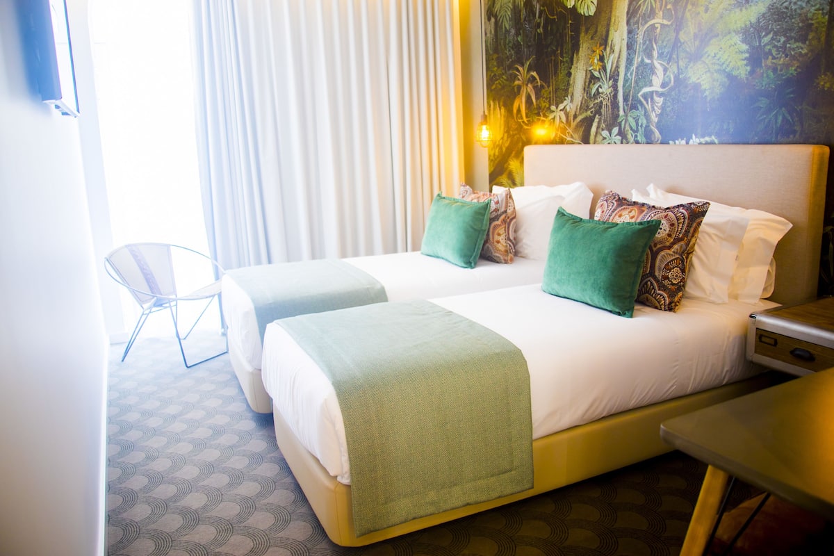 Ein Hotelzimmer mit zwei Betten, einem Sessel, einem Nachttisch und einem großen Gemälde an der Wand.