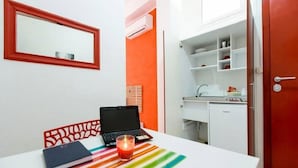 Una habitación pequeña con un escritorio blanco, una computadora portátil, un cuaderno y una vela. Hay un espejo en la pared y una puerta roja.
