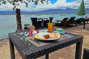 A dining table with a plate of food, a drink, and a cup, set against a scenic view of a body of water and mountains. - Salim Beach Resort (Gili Air)