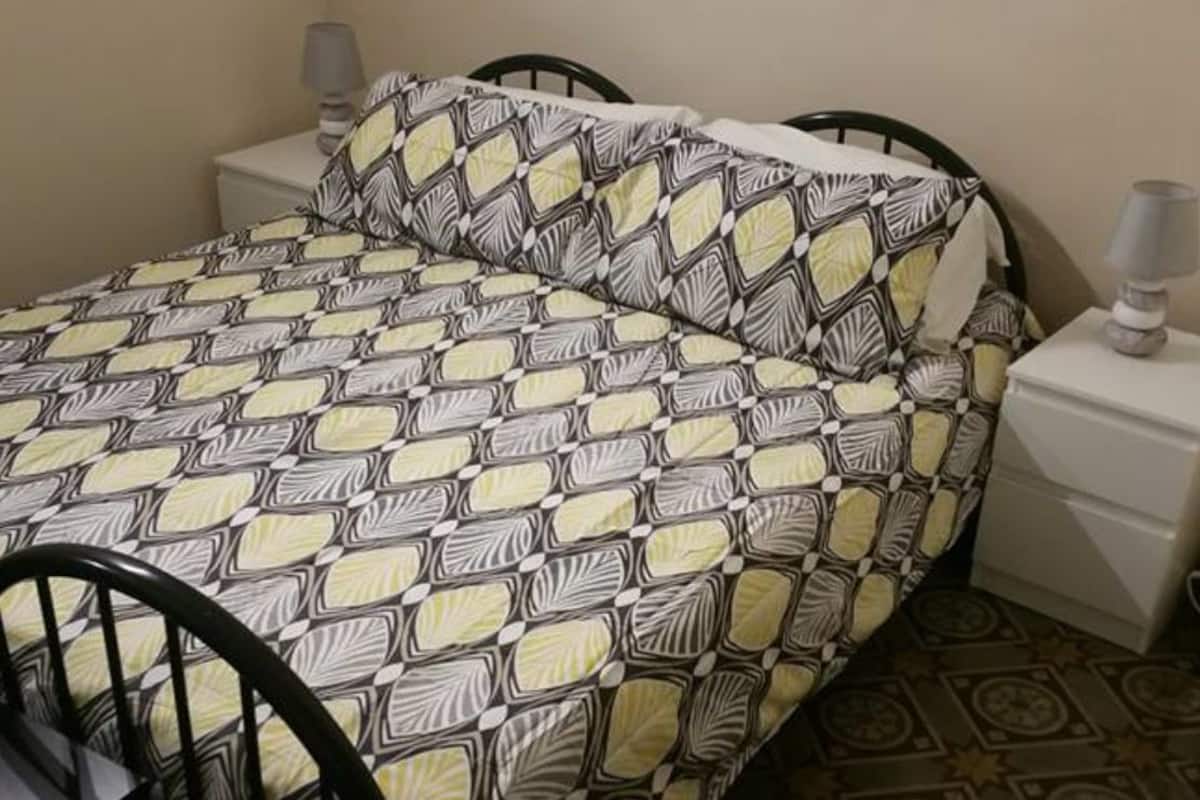 A bed with a patterned bedspread and pillows, a bedside table with a lamp, and a patterned floor.