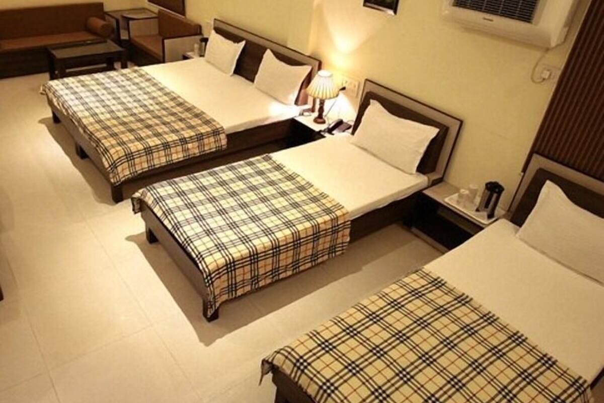 A hotel room with three single beds, each with a plaid bedspread, a bedside table, and a lamp. There are wall-mounted air conditioning units and a small picture on the wall.