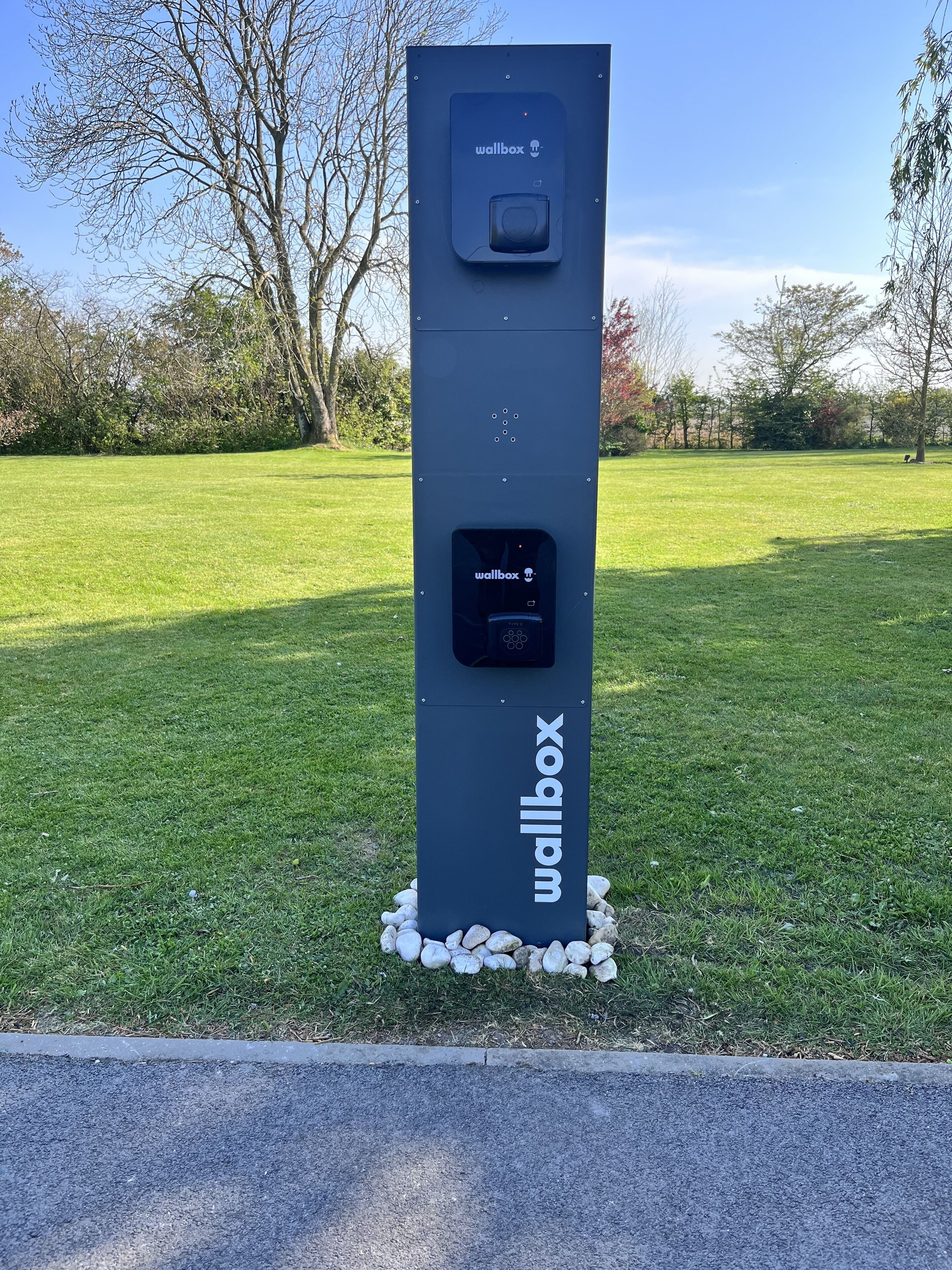 Een zwarte wandlader met het 'wallbox'-logo, gelegen op een grasveld met bomen op de achtergrond.