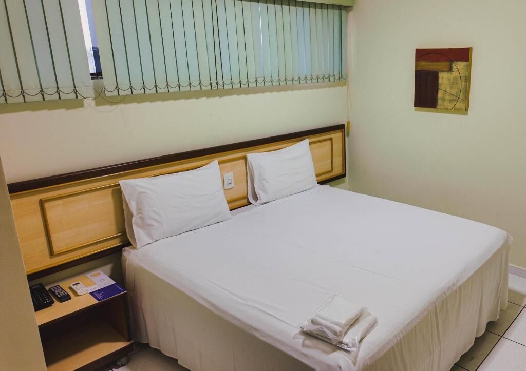 Habitació d'hotel amb un llit, una tauleta de nit, una finestra amb persianes i una obra d'art penjada a la paret.