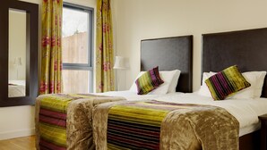 A double bed with a striped blanket and two pillows, a window with floral curtains, and a nightstand with a lamp. - Self Catering Holiday Homes Killarney Town Centre (Killarney)