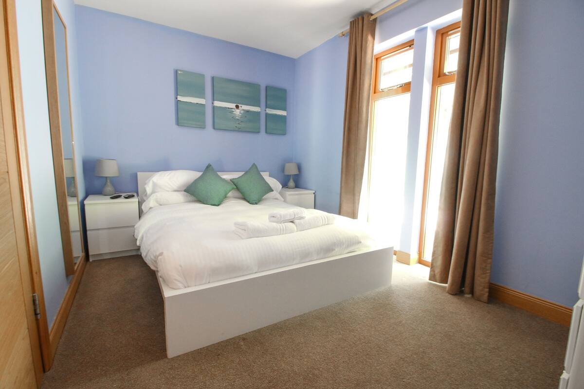 A neatly made bed with white linens, green pillows, and a folded towel. There are two bedside tables with lamps, and artwork on the wall.