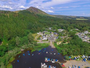 An aerial view of a lakeside village with boats docked at the marina, surrounded by dense forest and a prominent hill in the background. - Balmaha Inn (Drymen)