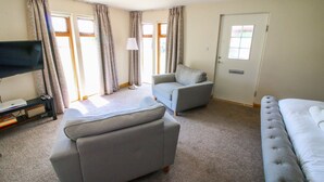 Un salón con un sofá gris, un sillón gris y una cama blanca. Hay un televisor sobre un mueble bajo y una lámpara sobre una mesa.