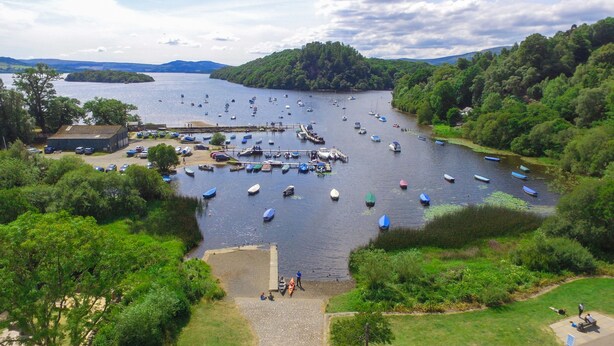 Vista del río con barcos amarrados a la orilla, un área verde con un sendero pavimentado y un edificio de techo plano.