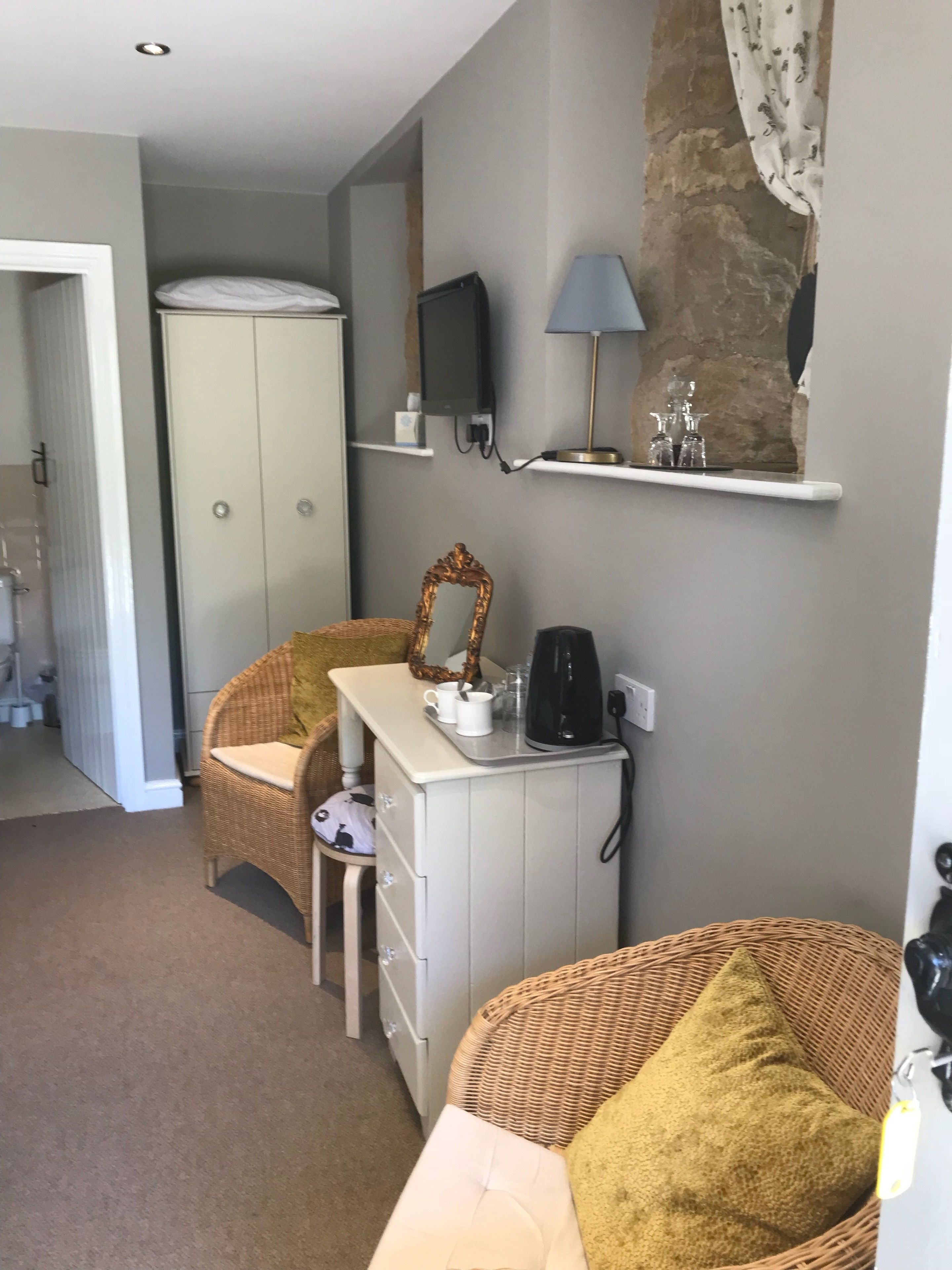 Une pièce de taille modeste, avec une armoire blanche, une chaise en osier, un petit bureau blanc doté d’un miroir, une lampe et une machine à café.