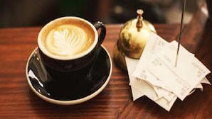 A cup of coffee with latte art on a saucer, a stack of receipts, and a small golden object on a wooden table. - Hotel Metro Jiapur (Jaipur)