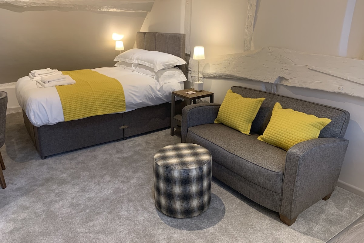 A bedroom with a bed, a grey sofa, a small ottoman, and two bedside lamps.