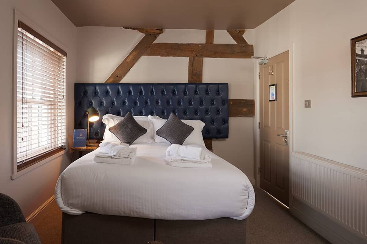 A bedroom with a large bed, a headboard, a window with blinds, a bedside lamp, and a door.