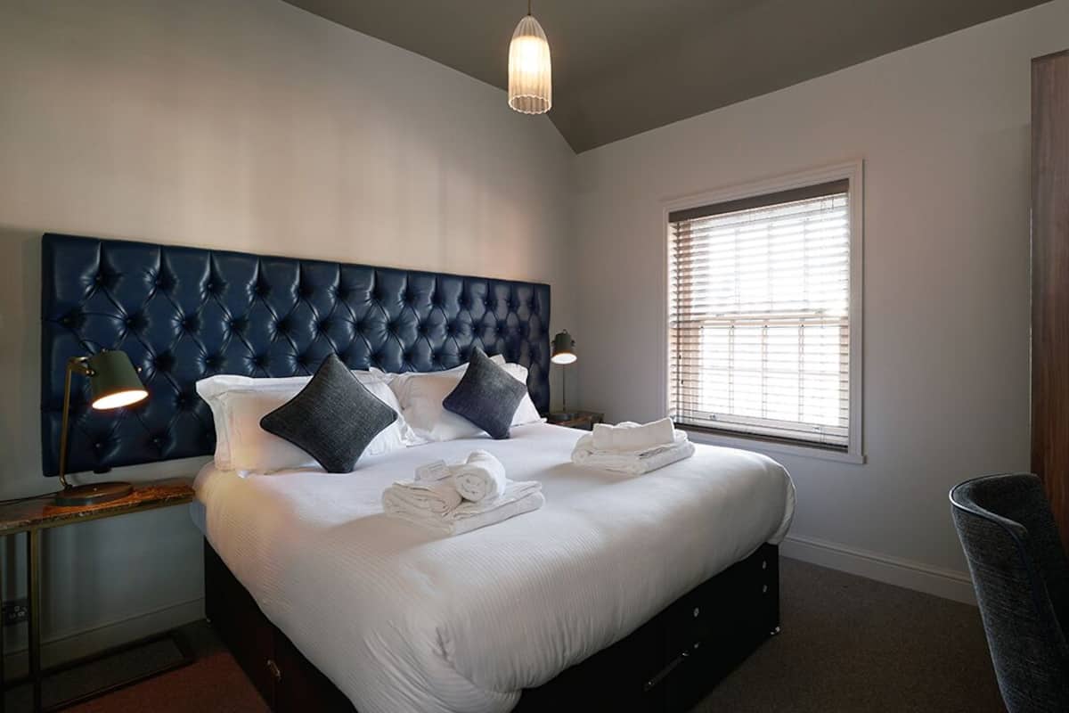 A bedroom with a large bed, a dark headboard, bedside lamps, a window with blinds, and a wooden wardrobe.