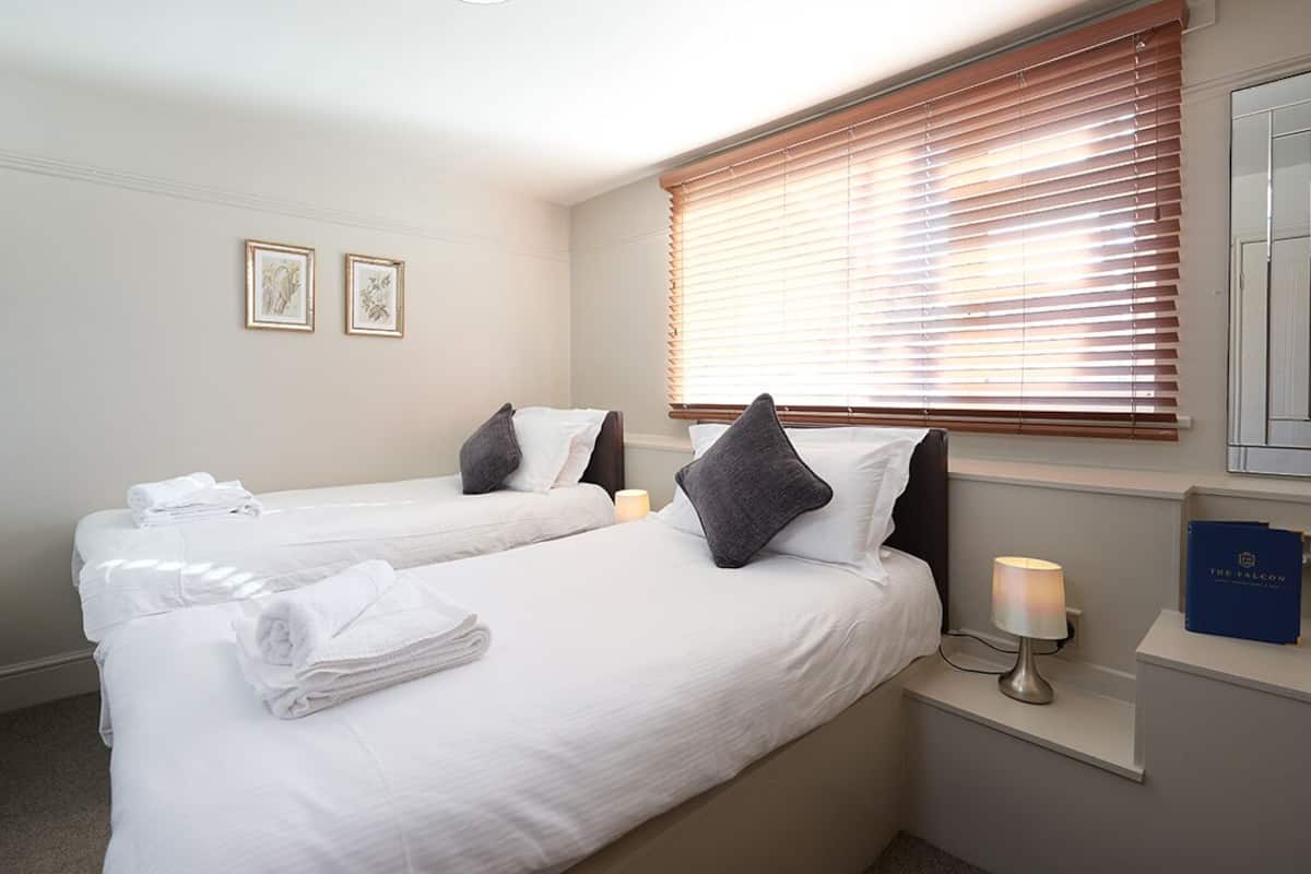 A hotel room with two beds, white linens, a bedside table with a lamp, a framed picture on the wall, and a window with blinds.