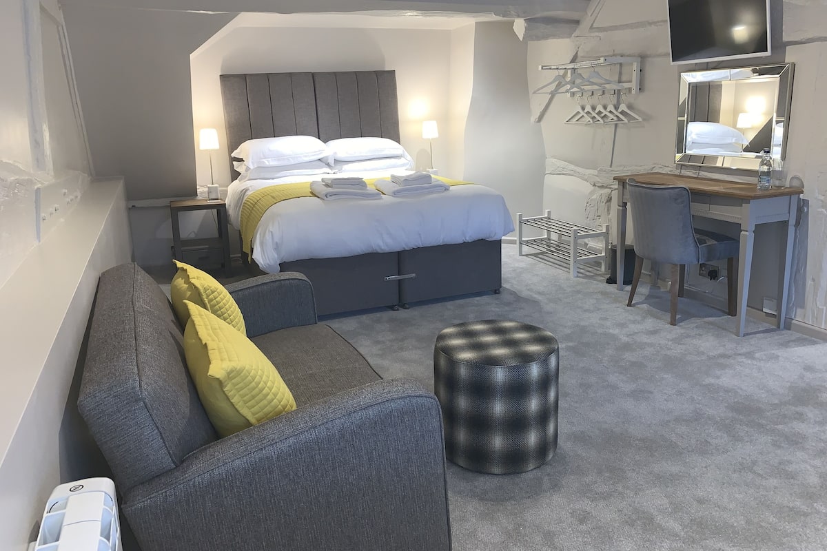 A modern hotel room with a bed, a desk, a chair, a sofa, and a small ottoman.