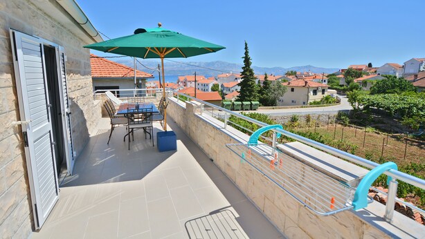 Un balcón con mesa y sillas, una sombrilla verde, y vistas a casas y montañas.