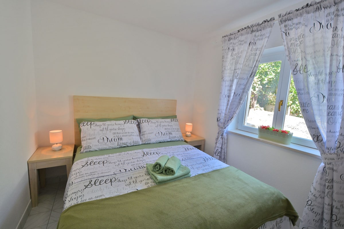 A neatly made bed with green bedding and pillows, a bedside lamp, and a window with curtains.