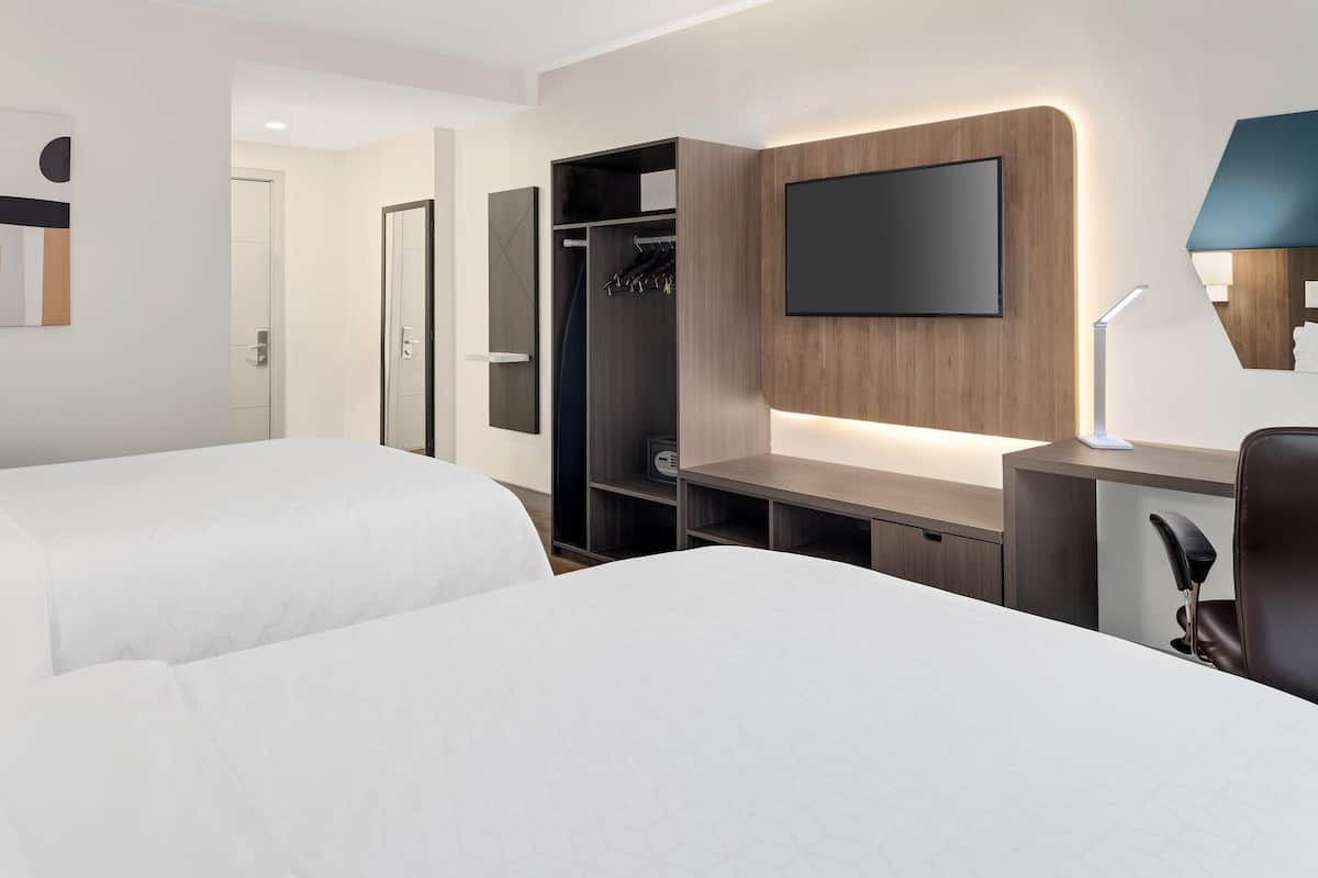 Habitación de hotel con dos camas, un televisor montado en la pared, un escritorio con lámpara y un armario.