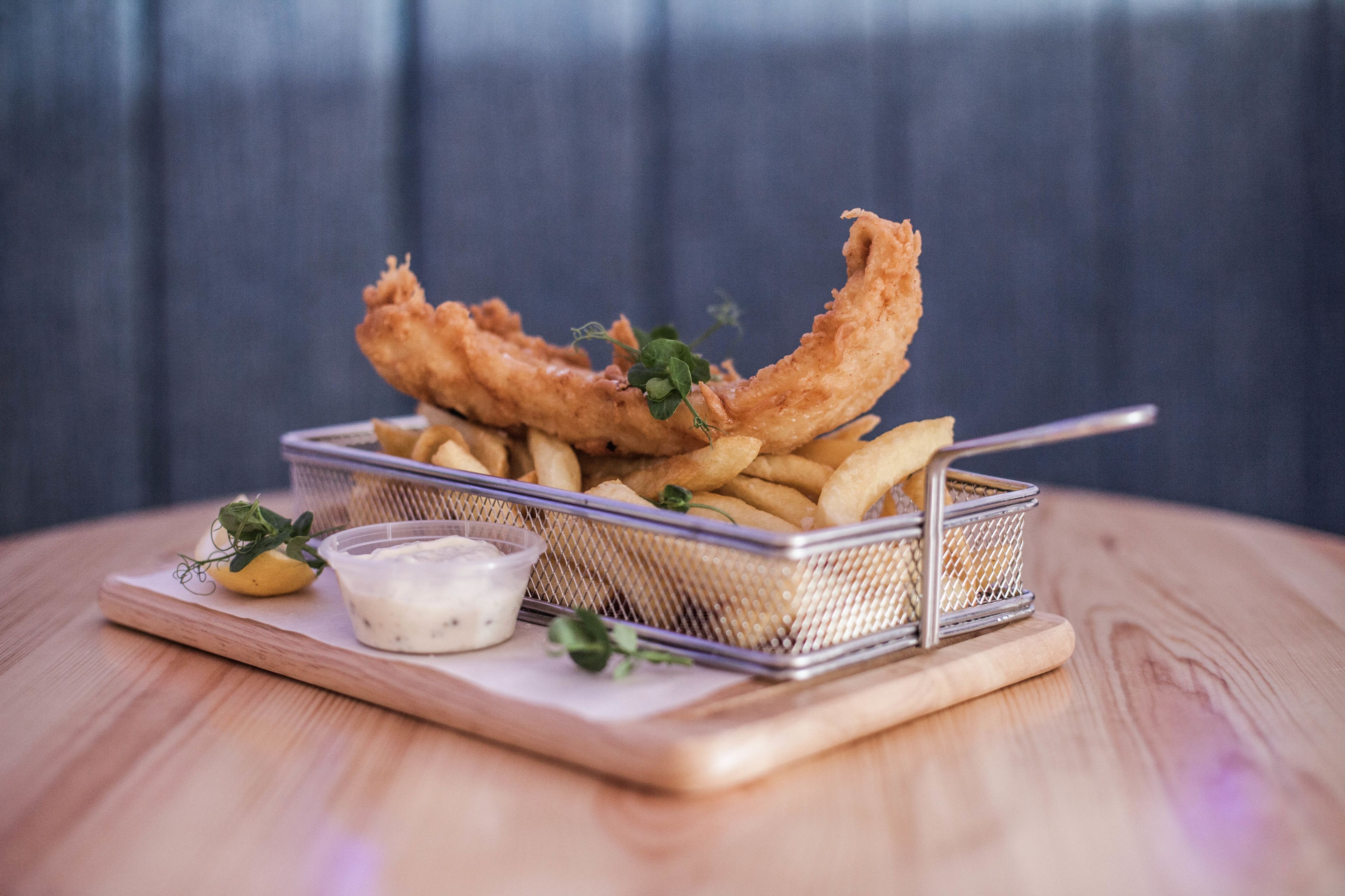 Una cesta de pescado frito y papas fritas sobre una tabla de madera, con un recipiente pequeño de salsa y un gajo de limón.