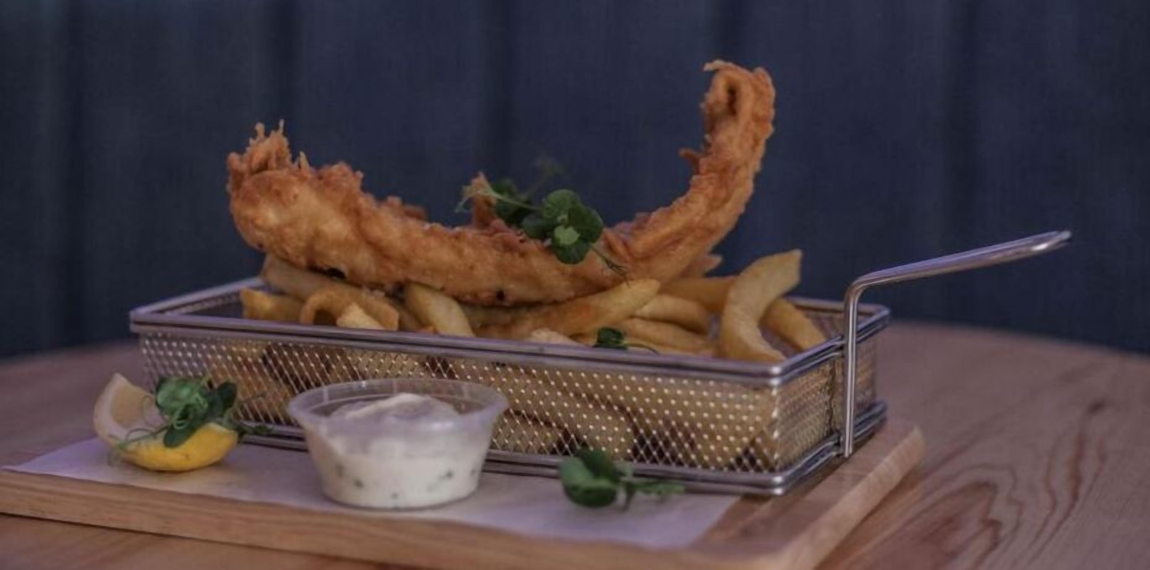 A basket of fried fish and chips with a small bowl of sauce and lemon wedges on a wooden table.